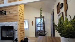A spacious living area with a wooden floor, a fireplace, a dining table, a hanging plant, and a black chair. - Hummingbird at Avenwood Cabins (New Straitsville)