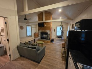 A spacious living area with a fireplace, a sofa, a television, and a kitchen area. - Hummingbird at Avenwood Cabins (New Straitsville)