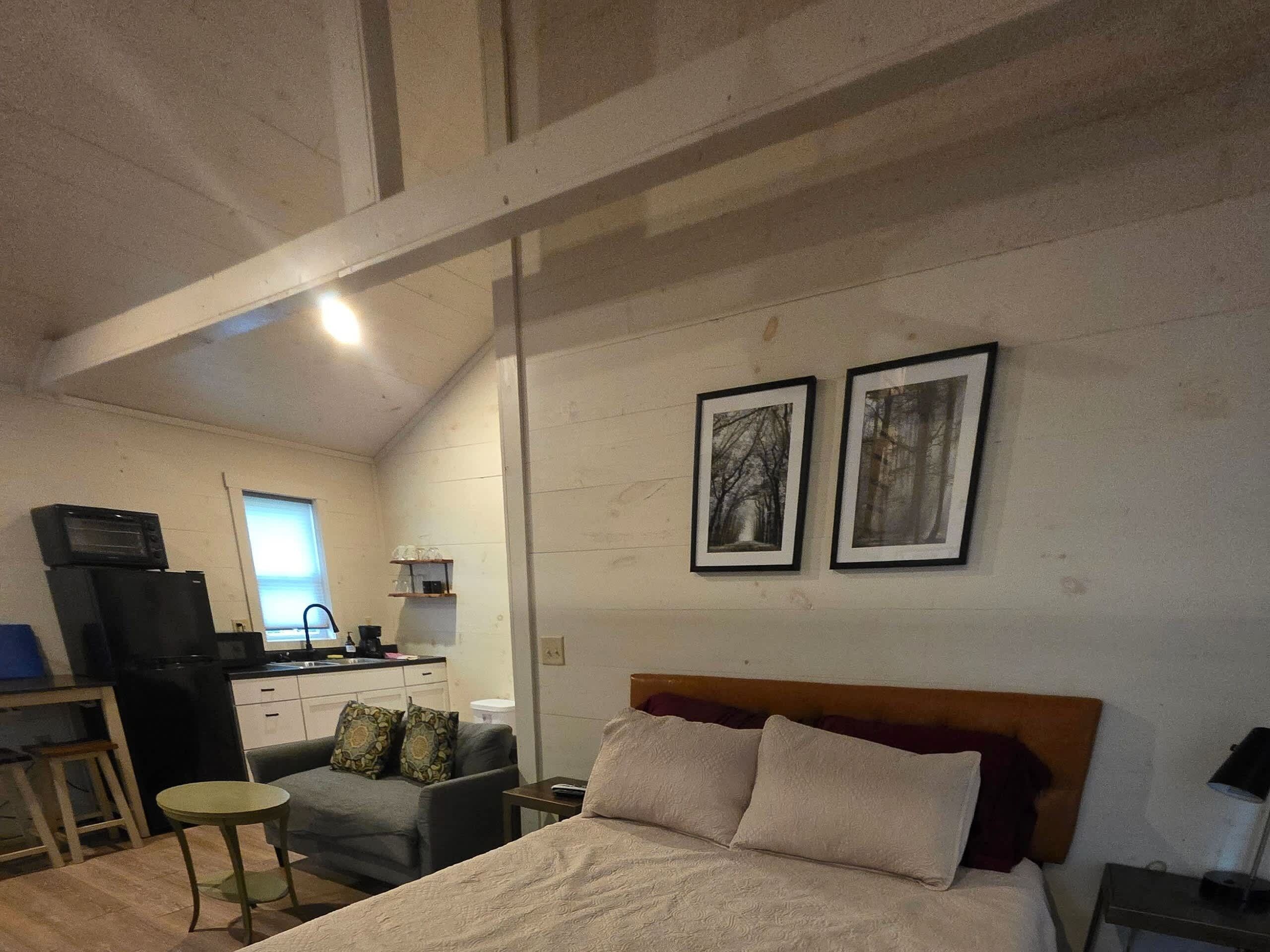 A bedroom with a bed, a desk, a chair, a small table, and a kitchenette in the background.