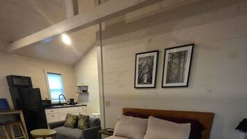 A bedroom with a bed, a desk, a chair, a small table, and a kitchenette in the background.