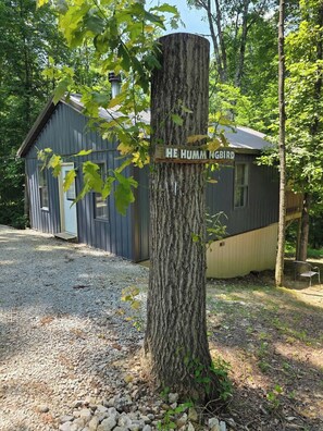 A rustic cabin with a wooden sign reading 'HE HUMM NGBIRD' on a tree. - Hummingbird at Avenwood Cabins (New Straitsville)