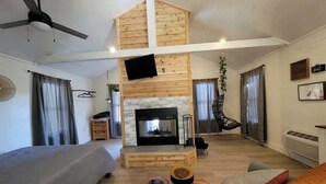 A spacious living area with a fireplace, a ceiling fan, and a lofted wooden structure. - Hummingbird at Avenwood Cabins (New Straitsville)