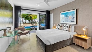 Une chambre moderne avec un grand lit, un bureau en verre, une chaise, une table de chevet avec une lampe, et une vue sur une piscine et la verdure environnante.