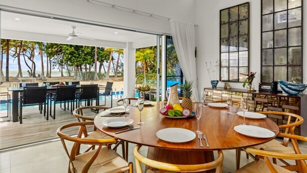 Un coin repas avec une table en bois dressée pour six personnes, entourée de chaises en osier, donnant sur une piscine et des palmiers.