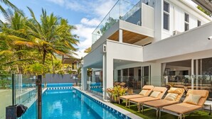 Une maison moderne dotée d’une piscine, de chaises longues et d’un balcon fermé par des baies vitrées.