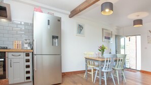 A kitchen with a stainless steel refrigerator, a water dispenser, and a coffee machine. There is a wooden dining table with chairs and a vase of flowers. - Old Highwood - Piglet Cottage (Dunkeswell)