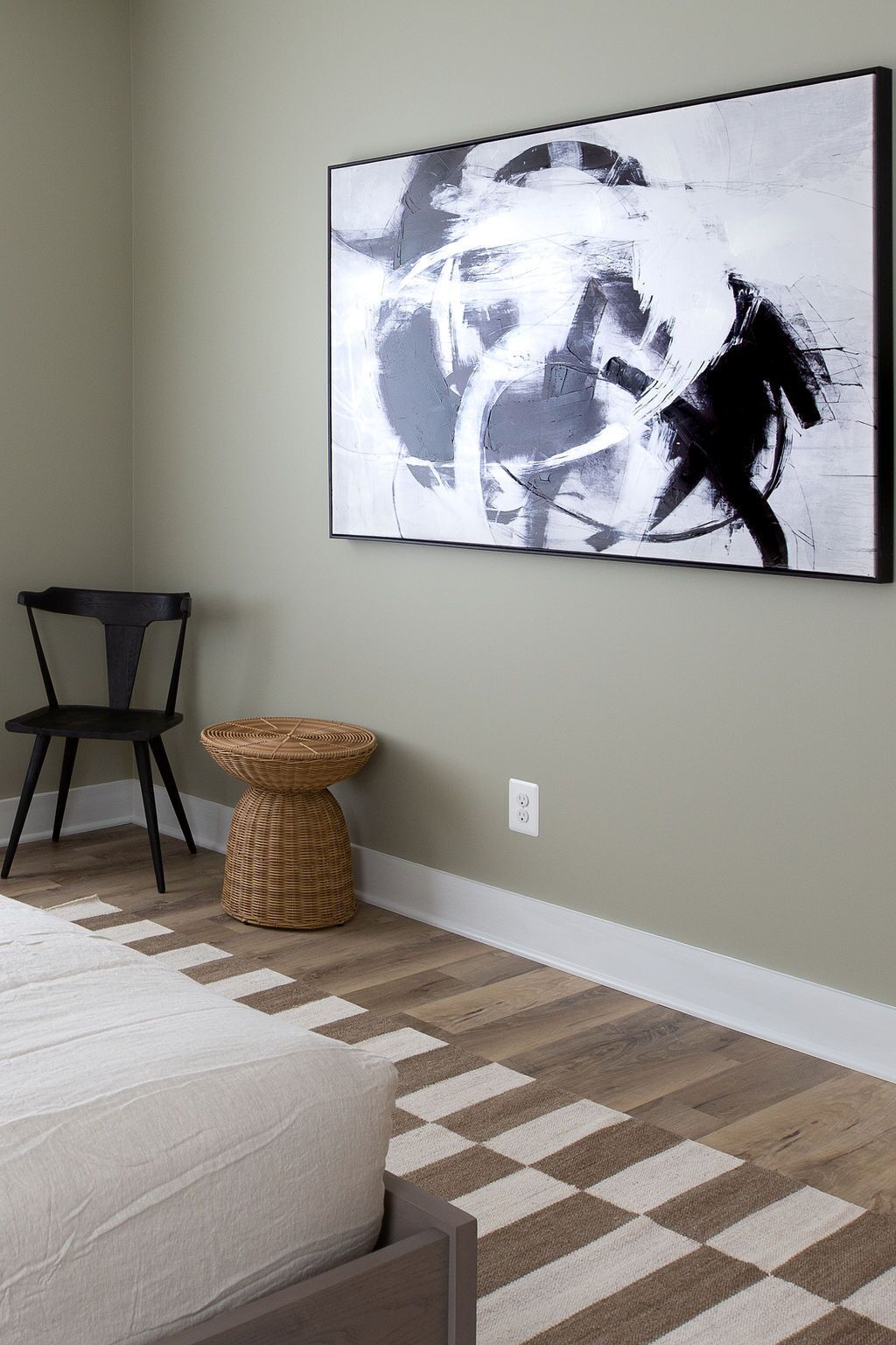 A bedroom with a bed, a chair, a woven stool, and a large abstract painting on the wall.