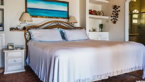Cama bem arrumada com colcha branca e almofadas, cabeceira de madeira, mesas de cabeceira com abajures, prateleira com objetos decorativos e uma pintura de uma praia.