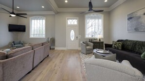 A spacious living room with a large sofa, a smaller armchair, and a dark-colored couch. There are two windows with decorative glass panes, a ceiling fan, and a flat-screen TV mounted on the wall. - The Nashville Oasis | Ultimate Outdoor Escape (Nashville)
