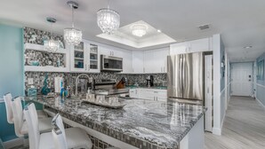 Cocina moderna con armarios blancos, encimeras de granito y electrodomésticos de acero inoxidable. Hay dos lámparas colgantes y un tragaluz sobre la isla.