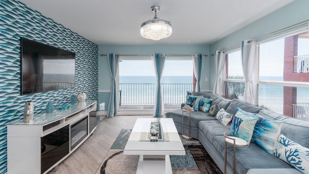 Una sala de estar moderna con un televisor grande, una mesa de centro blanca y un sofá gris con cojines azules. Hay un balcón con vista al mar.