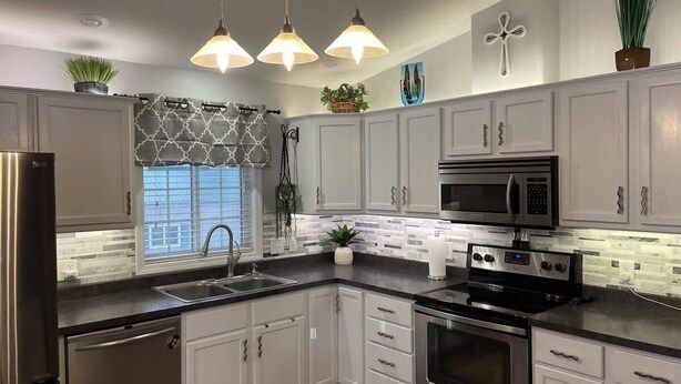 Eine Küche mit weißen Schränken, einem Edelstahlkühlschrank, einer Mikrowelle, einem Backofen und einer Spüle. Drei Pendelleuchten, ein Fenster mit einem gemusterten Vorhang und ein Blumentopf auf der Arbeitsplatte.