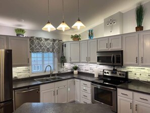 A kitchen with white cabinets, a stainless steel refrigerator, a microwave, an oven, and a sink. There are three pendant lights, a window with a patterned curtain, and a potted plant on the counter.