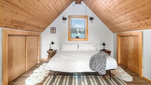 A bedroom with a sloped ceiling, a bed with white bedding, a window with blinds, and wooden doors.