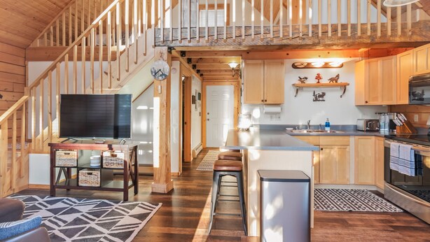 A spacious loft-style living area with a wooden staircase, a large flat-screen TV, a dining table, and a kitchen with wooden cabinets and a stainless steel range hood. - Dreamy 2 Bed Cabin #2 - Alaska Kenai Getaway (Sterling)