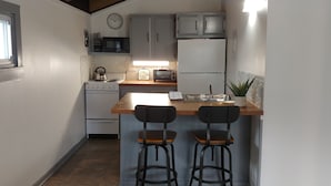 A compact kitchen with a stove, microwave, refrigerator, and a small bar area with two stools. - Binnekill Views- Cozy Apt over looking the Binnekill stream (Margaretville)