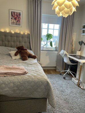 A bedroom with a bed, a desk, a chair, a window with curtains, a plant, and a hanging light fixture.