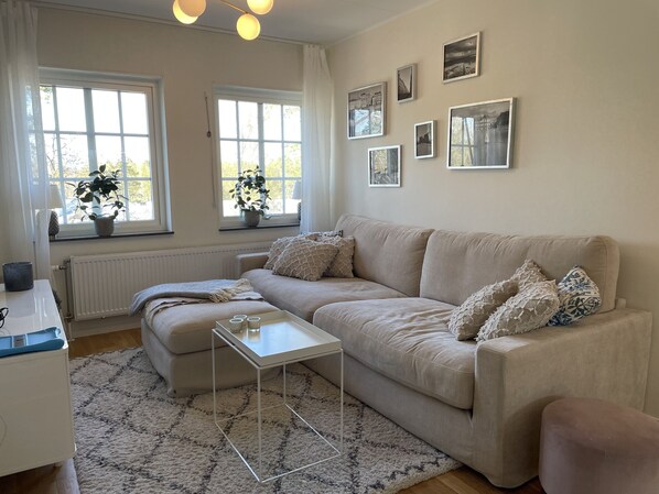 A living room with a beige sofa, a white coffee table, and a patterned rug. There are two windows with curtains, a radiator, and several framed pictures on the wall.
