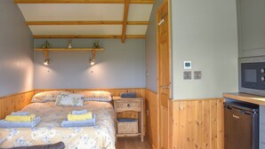 A bedroom with a wooden ceiling, a bed with floral bedding, a bedside table, a small shelf with decorative items, and a built-in microwave.