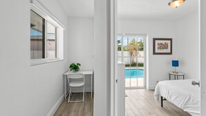 A bedroom with a bed, a desk, a chair, a plant, a framed picture, and a view of a swimming pool.
