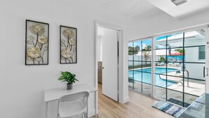 A modern room with a glass door leading to a pool area, a white desk with a chair, and two framed floral artworks on the wall.