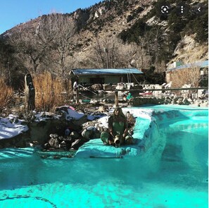 A natural hot spring with clear turquoise water, surrounded by rocks and sparse vegetation.