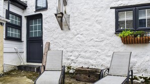 Une cour avec deux chaises en métal, une porte noire et une fenêtre agrémentée d’une jardinière.
