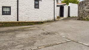 Une allée étroite, recouverte de gravier, mène à un bâtiment blanc à la façade texturée, doté d’une porte noire et d’une petite fenêtre.