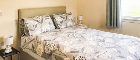 A neatly made bed with patterned bedding, a bedside table with a lamp, and a window with curtains.