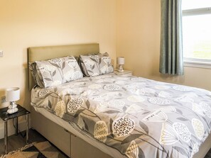 Una cama bien tendida con ropa de cama estampada, una mesita de noche con una lámpara y una ventana con cortinas.
