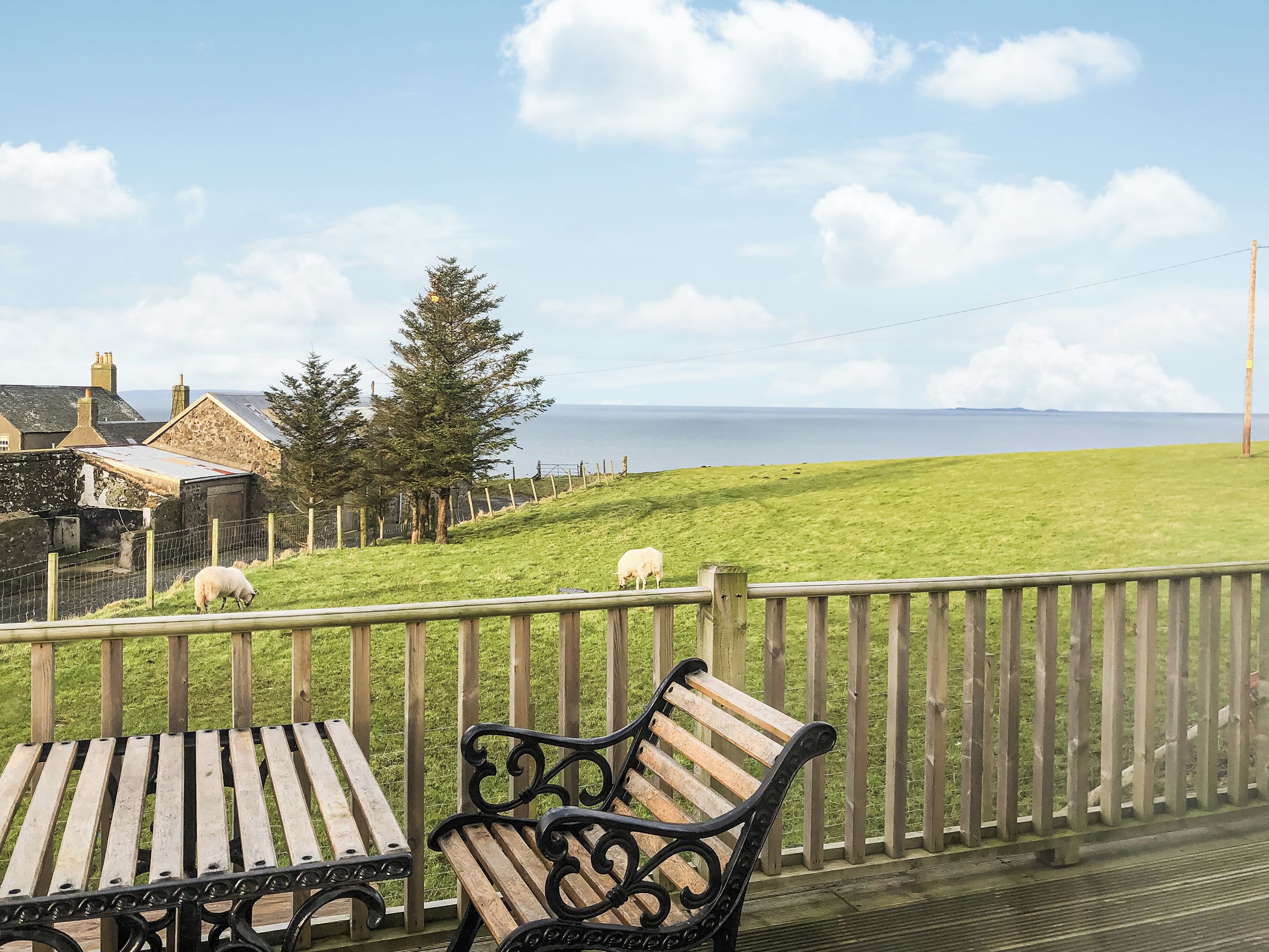 Une terrasse en bois avec des bancs, donnant sur une zone herbeuse où paissent des moutons, une clôture et une vue sur la mer.
