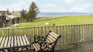 Une terrasse en bois avec des bancs, donnant sur une zone herbeuse où paissent des moutons, une clôture et une vue sur la mer.