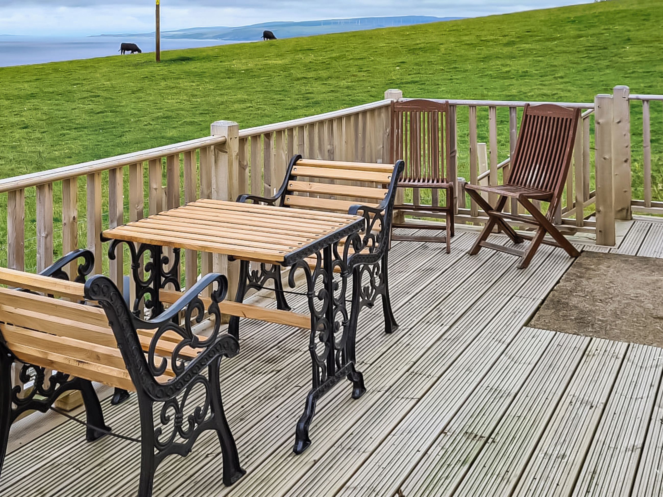 Eine Holzterrasse mit Gartenmöbeln, darunter Bänke und ein Tisch, mit Blick auf eine Rasenfläche und das Wasser.