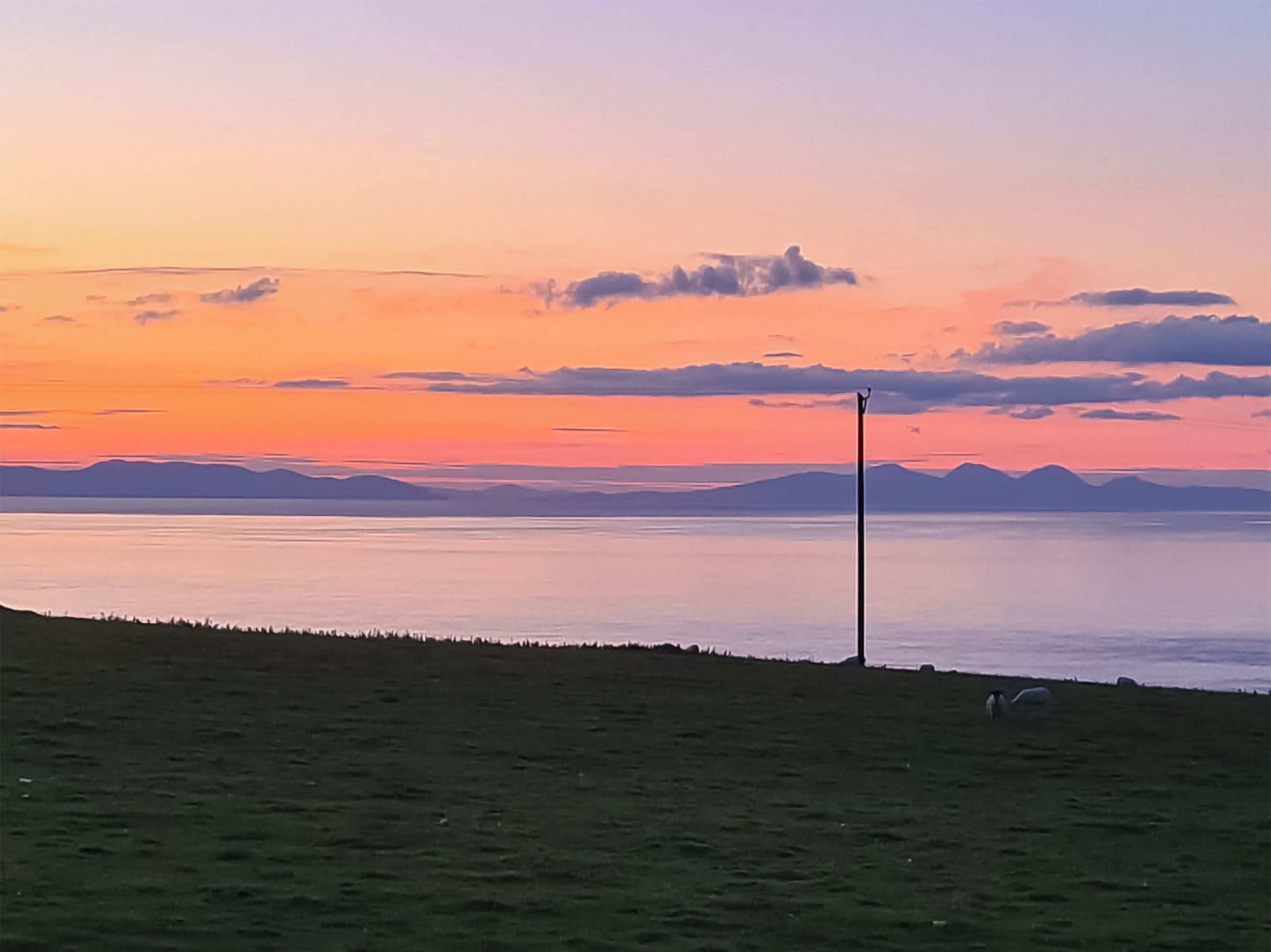 A serene landscape with a body of water, distant mountains, and a grassy area with a few sheep.