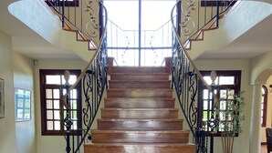 Una imponente escalera de madera con barandales negros ornamentados, que conduce a un espacio interior luminoso con amplios ventanales y una planta en maceta.