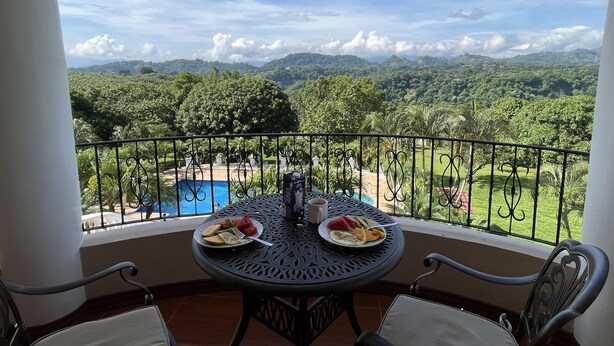 Un balcón con una mesa preparada para dos, con vistas a un paisaje exuberante y una piscina.