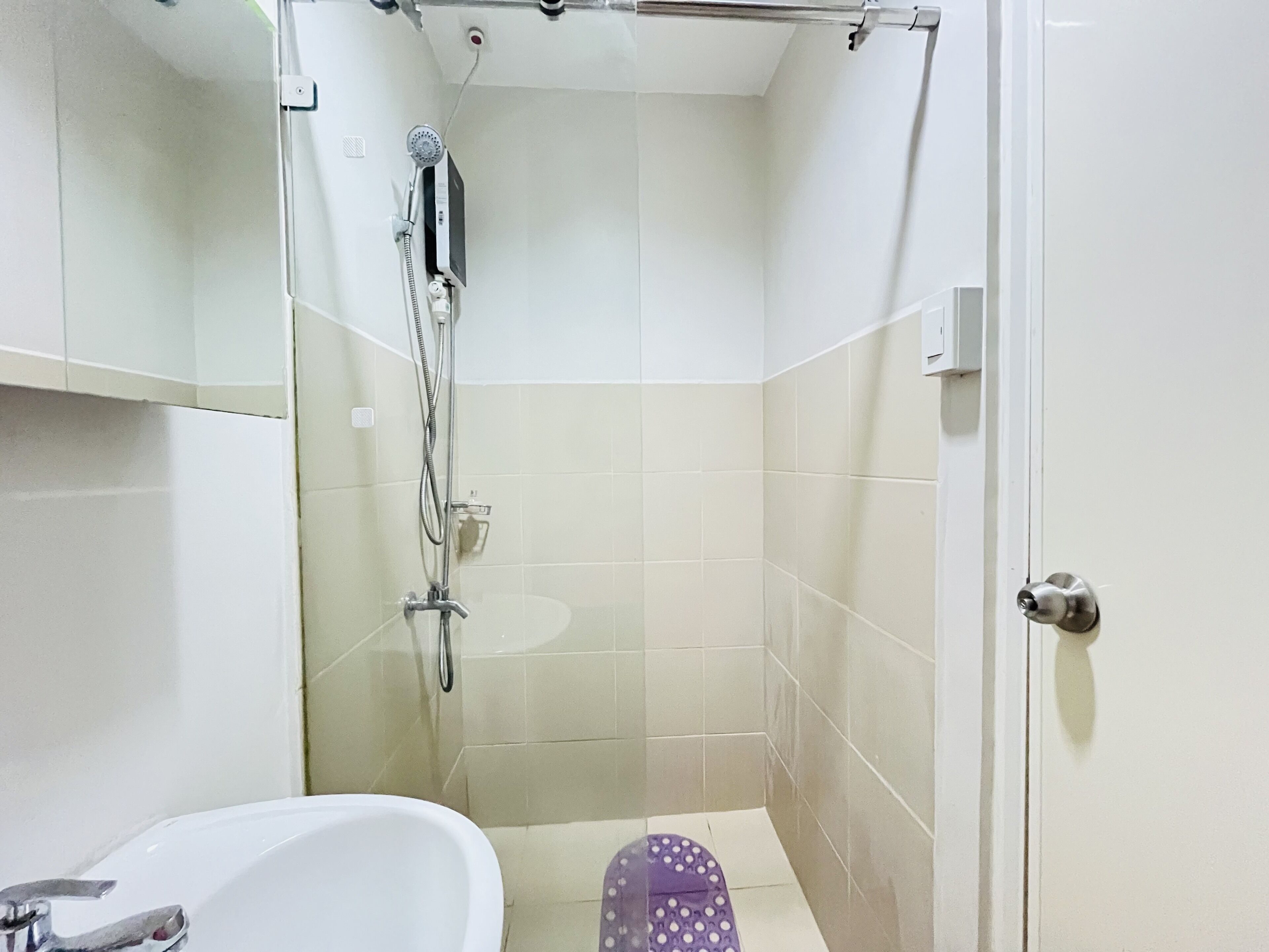 Un petit espace douche fermé, doté d’un pommeau de douche à main, d’une baignoire blanche et de toilettes violettes.