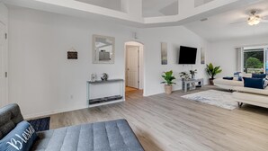 A spacious living room with a gray sofa, a coffee table, and a TV mounted on the wall. There are potted plants and a wooden floor. - **EXCLUSIVE 3-bedroom w/heated private pool** (Palm Coast)