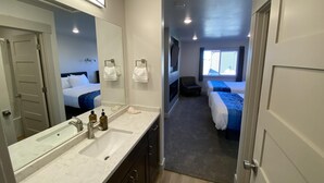 Una habitación de hotel moderna con un espejo grande, un lavamanos con dispensadores de jabón, una silla y una cama con una manta azul.