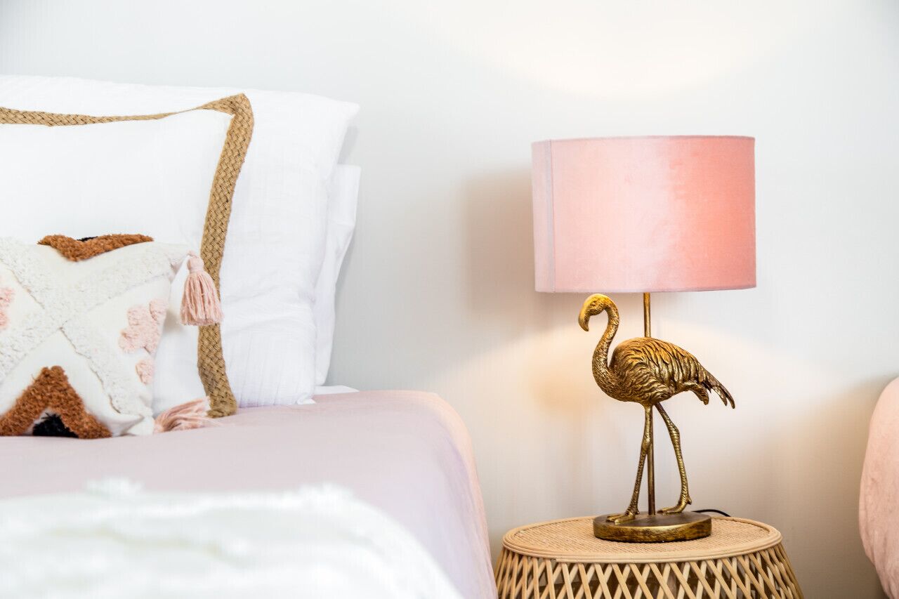 Uma cama com um cobertor rosa, um travesseiro branco e um travesseiro decorativo com padrão marrom e rosa. Há uma luminária com cúpula rosa e um flamingo dourado sobre uma mesa de madeira redonda ao lado da cama.