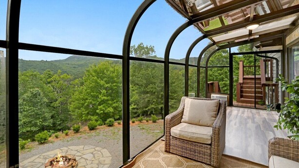A glass-enclosed patio with a wicker sofa and a fire pit, overlooking a lush landscape. - Ridge View - Hot Tub-Fireplace-Fire Pit (Hiawassee)
