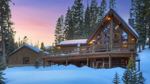 Una cabaña de troncos con balcón y ventanas amplias, rodeada de nieve y árboles.