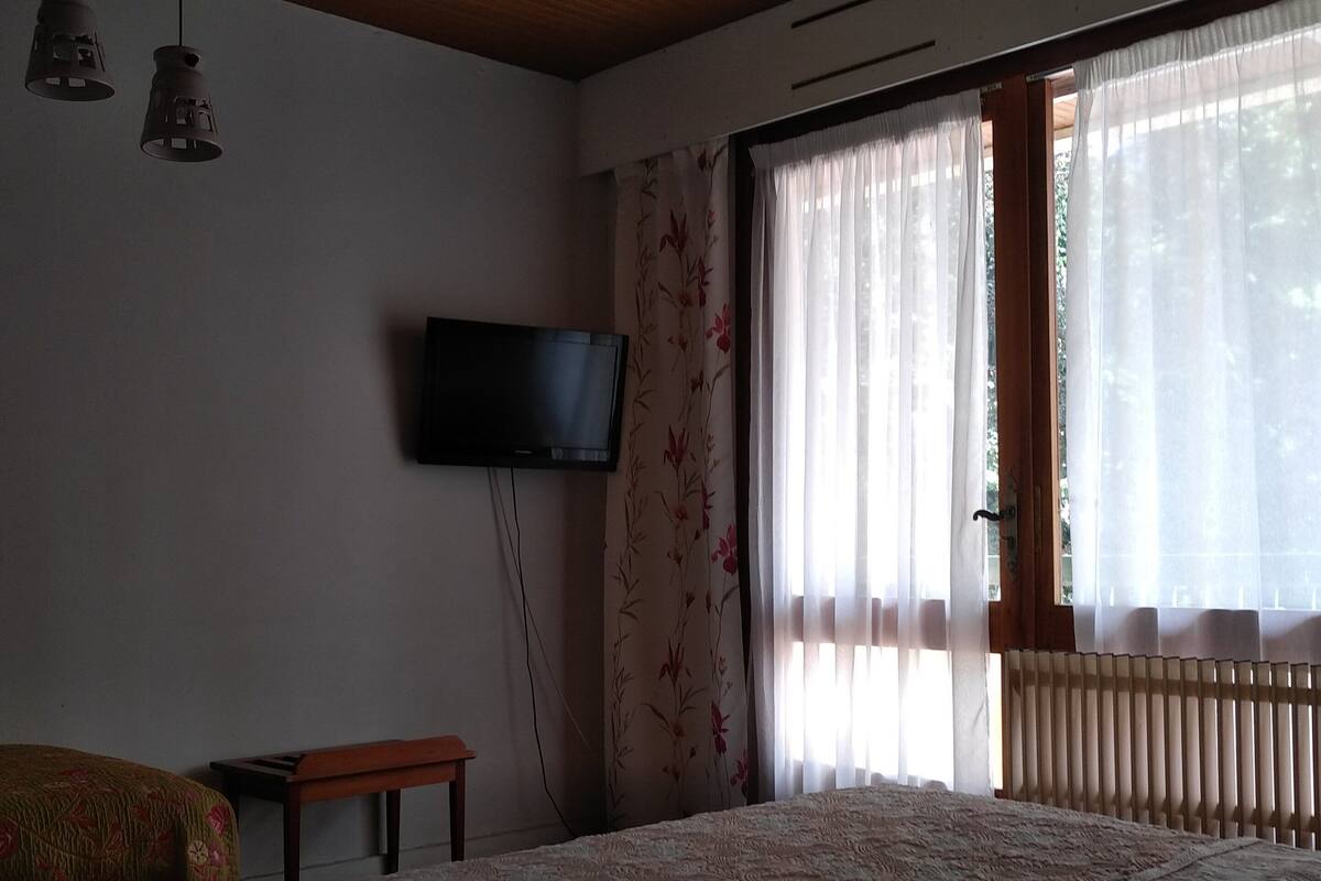 Une chambre avec un grand lit, un téléviseur fixé au mur, un banc et une fenêtre avec des rideaux.