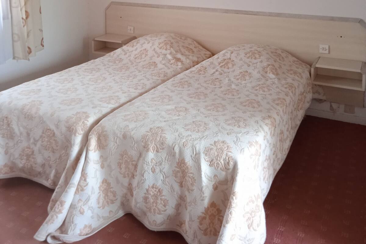Une chambre avec un plafond en bois, deux lits simples avec une literie à motifs floraux, une petite étagère et un rideau.