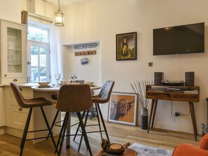 Ein Essbereich mit Tisch für vier Personen, eine Küche mit Glaskabinett und ein an der Wand montierter Flachbildfernseher.