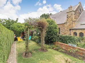 A garden with a pergola, a brick wall, and a stone building with a tiled roof.
