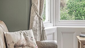 Une pièce comprenant un canapé, un coussin à motifs floraux, une fenêtre avec des rideaux et une petite table en bois sur laquelle se trouve un livre.