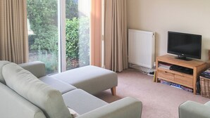 Un salon avec un canapé, un téléviseur posé sur un meuble en bois, et une vue sur la verdure à l’extérieur.