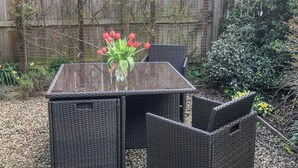 Une table en osier ornée d’un vase de fleurs rouges, placée devant une clôture en bois et sur fond de jardin.