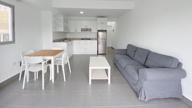 A modern living room with a grey sofa, white dining table, and chairs, a white coffee table, and a kitchen area with white cabinets and appliances. - Your ideal vacation home in Sosua! (Sosúa)